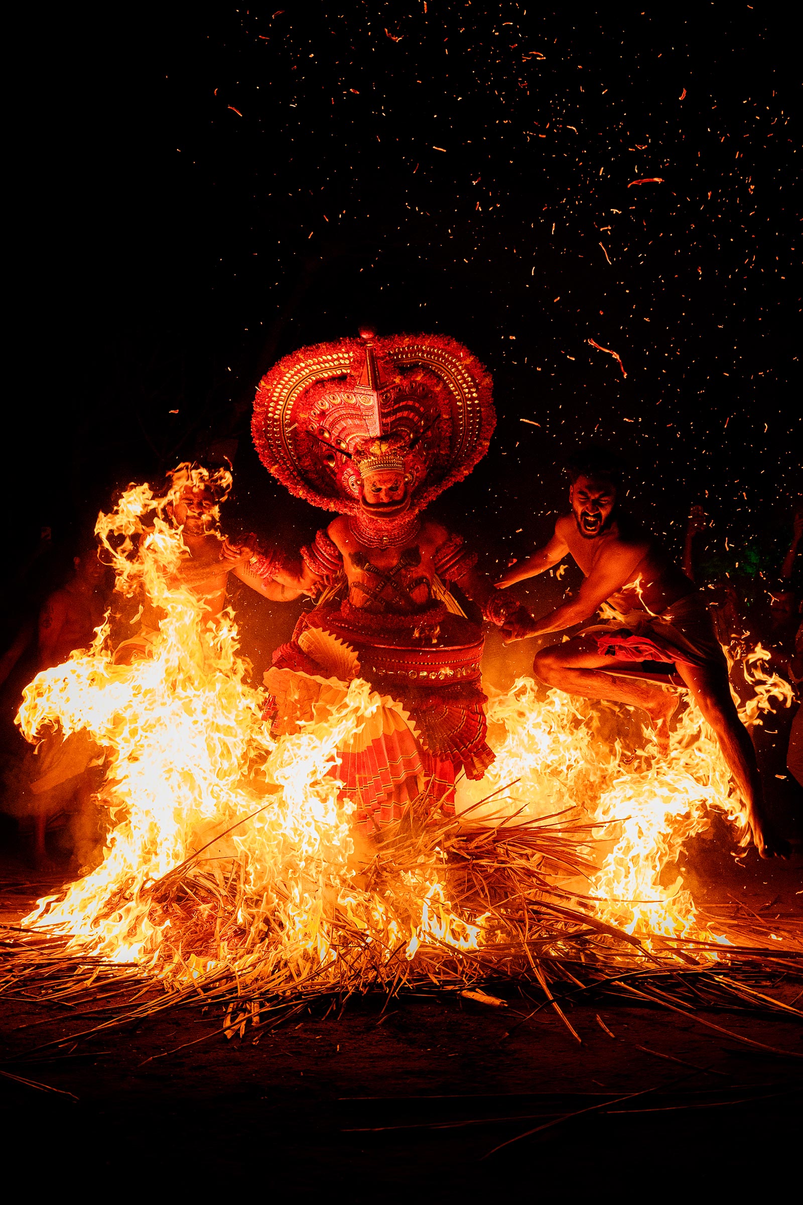 The image depicts a vibrant scene from a the fire-act of Kandanar Kelan Theyyam. Here, a central figure, adorned in elaborate red costume and a large headdress, jumps above a roaring bonfire. Two other individuals, seemingly bare-chested and participating in the ritual, leaps across the flames. The overall impression is one of intense energy, spectacle, and of cultural significance. The dark background and sparks emphasize the dramatic lighting and action.